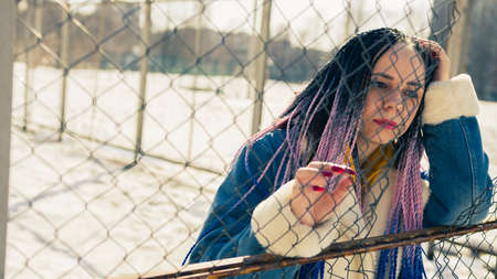 Serious young female in warm clothes looking away while leaning on net fence on city street