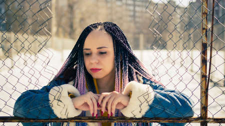 Serious young female in warm clothes looking away while leaning on net fence on city street