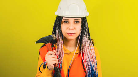 Female construction worker in overalls with hammer on yellow background in studio. Young woman in...