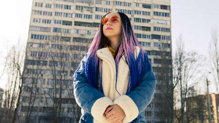 Low angle side view of young female in warm clothes and trendy sunglasses standing on street and...