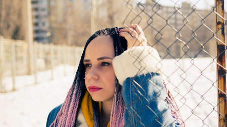 Serious young female in warm clothes looking away while leaning on net fence on city street