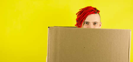Young man looking out of cardboard box on yellow background in studio. Male courier hiding behind...
