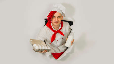Man with red dreadlocks holds it in his hand chicken eggs and milk. Confident young cook in white apron and red tie holding chicken eggs and a bottle of milk, peeking through ripped paper background.の写真素材