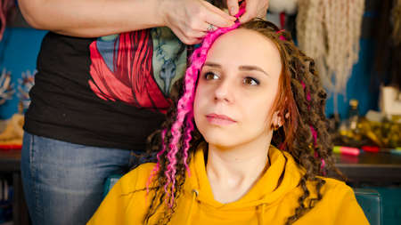 Unrecognizable person makes curly hairstyle for young woman in salon. Professional hairstylist makes pink and brown dreadlocks for clientの写真素材