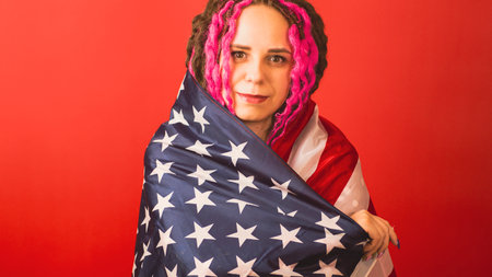 Young woman wrapped in American flag on red background. Happy female smiling and looking at camera, posing in studio. Concept of Flag Day, national holidayの写真素材