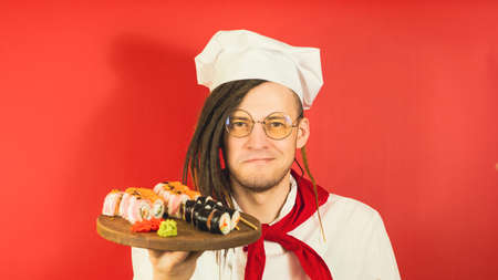 Young man in glasses dressed as chef holding wooden board with sushi rolls. Male cook with appetizing sushi rolls on red backgroundの写真素材