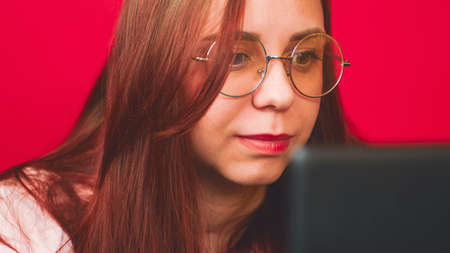 Young woman using laptop in studio. Positive young female in eyeglasses and casual clothes smiling and looking watching videos on the computer in modern office against red background.の写真素材