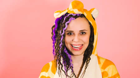 Young cute woman with dreadlocks in kigurumi looking at camera and showing tongue. Funny playful female with curly hairstyle in pajamas on pink backgroundの写真素材