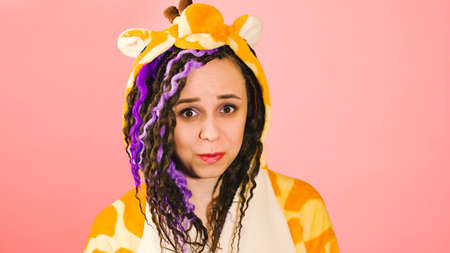 Young shy beautiful woman in kigurumi looking at camera on pink background. Cute vivid female with dreadlocks in pajamas smiling with confused facial expressionの写真素材