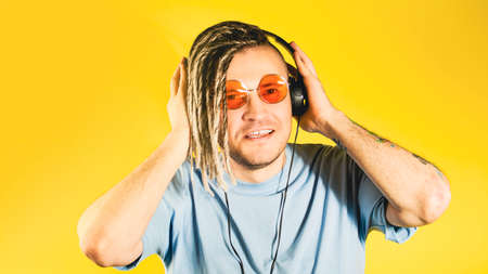 Young handsome man in sunglasses, headphones listening to music, singing. Positive cheerful guy with blonde dreadlocks dancing, enjoying songs through headphones on isolated yellow backgroundの写真素材