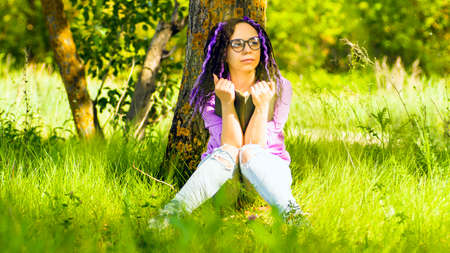 A woman in a purple shirt and glasses reads a literature under a tree. A girl is resting with a book...