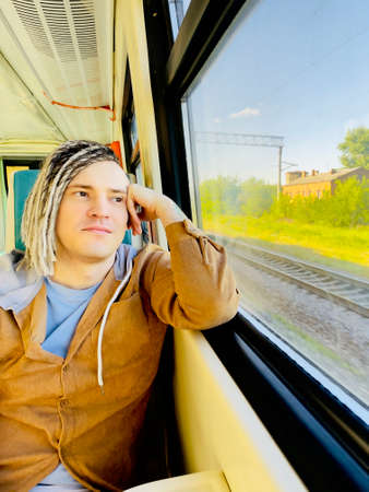 Young relaxed man riding on train. Dreamy guy with blonde dreadlocks looking at nature through...