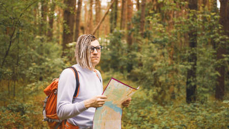 Traveling man with dreadlocks with map in woods. Male traveler with backpack in woods reading map in...