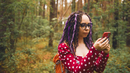 Traveling woman with dreadlocks with a phone in woods. Female traveler with backpack in woods reading map on a smartphone and looking for a way in cloudy weather.の写真素材