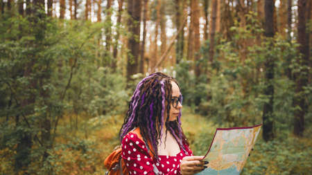 Traveling woman with dreadlocks with map in woods. Female traveler with backpack in woods reading...