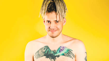 Naked handsome young man posing with positive emotion. Attractive guy with blonde dreadlocks, tattoo on chest looking at camera, smiling on yellow backgroundの写真素材