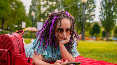 A girl is watching a video on her phone, lying on a red blanket in a park in summer. A woman in a dress watches content on a smartphone in a city park.の写真素材