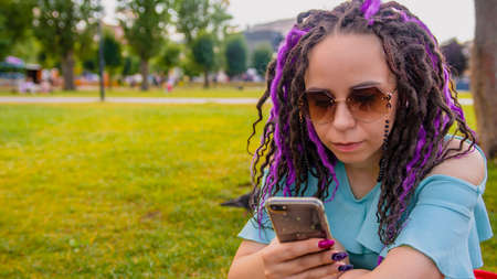 Young woman sitting on a blanket in the park and using a smartphone. Happy young brunette sitting on...
