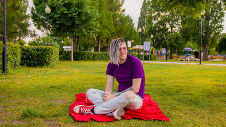 A handsome guy with dreadlocks in casual clothes, sitting on a plaid in a park on a green lawn and looking thoughtfully into the distance. A handsome man is sitting on the grass on a sunny summer dayの写真素材