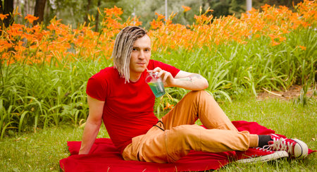 Young man drinking refreshing lemonade, sitting on rug in city park. Relaxed male with dreadlocks drinking soft drink through straw, quenching thirst, resting on lawn on background of blurred flowersの写真素材