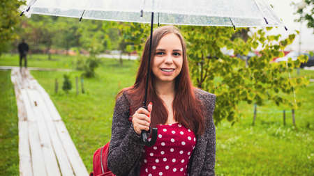 Young happy beautiful woman with transparent umbrella in rain, standing in city park. Portrait of...