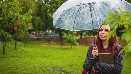 Young beautiful woman with transparent umbrella hugging book, standing in city park in rain. Dreamy...