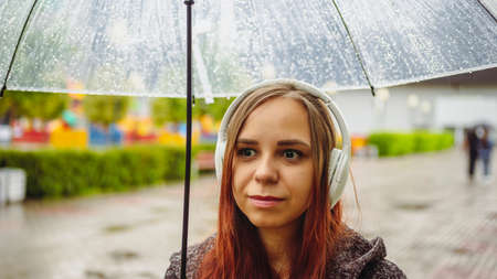 Young beautiful woman in headphones listening to music, standing with transparent umbrella in rain...