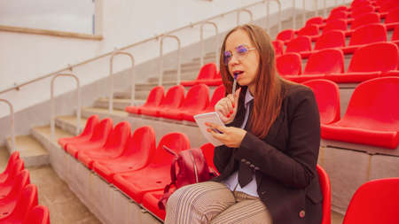 Young woman in glasses with notepad, pen sitting on stadium bleachers alone. Female journalist...