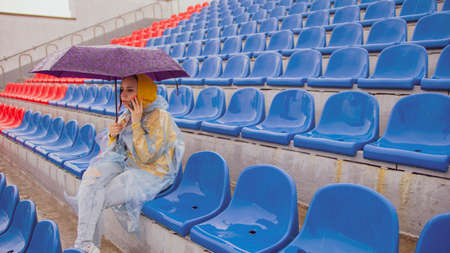 A beautiful woman in a hoodie and a plastic raincoat, talking on a mobile phone, sitting on the...
