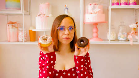 A beautiful woman in a red dress and glasses holds a white donut in one hand and a chocolate donut...