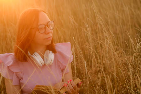 Thoughtful young female in casual clothes and eyeglasses sitting in dry grass and reading a book at sunset.の写真素材