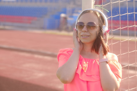 A beautiful woman in an orange dress and sunglasses listens to music using white headphones at the stadium, leaning on a football goal.の写真素材