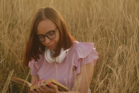 Thoughtful young female in casual clothes and eyeglasses sitting in dry grass and reading a book at sunset.の写真素材