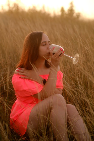 Young woman with a glass of wine in the field. Side view of young female in orange dress drinking red wine and looking away while sitting on grass in countryside.の写真素材