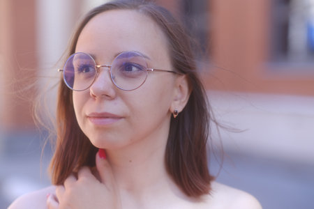 Portrait of young woman with short hair in glasses looking away, standing on city street in warm windy weatherの写真素材
