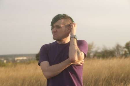 Young thoughtful man with green dreadlocks with doubt glance scratching head in countryside on eve of sunsetの写真素材