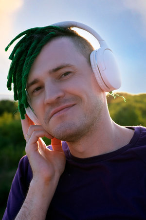 Portrait of young happy man with green dreadlocks, wireless headphones listening to music in nature...