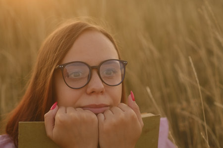 Young dreamy woman in glasses hugging book, sitting in dry grassの写真素材