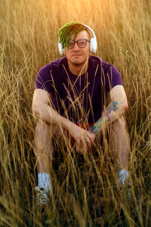 Young handsome man in glasses with green dreadlocks, wireless headphones listening to music in...
