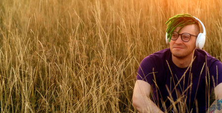 Young handsome man in glasses with green dreadlocks, wireless headphones listening to music in nature, sitting in dry grassの写真素材
