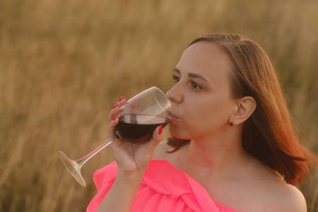 Young woman in bright dress with short hair drinking red wine, standing in nature on sunsetの写真素材