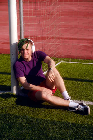Young man in wireless headphones listening to music, closing eyes from bright sun, sitting on football field, leaning on gatesの写真素材