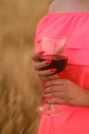 Unrecognizable woman holding glass of red wine, standing in nature on sunset. Body part of female in bright dress with alcohol in countrysideの写真素材