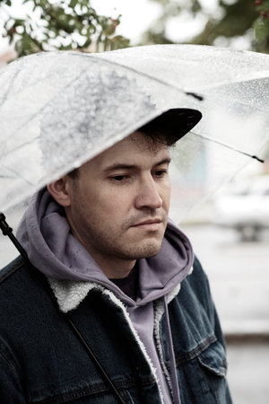 Young pensive man standing with transparent umbrella in rain on city street