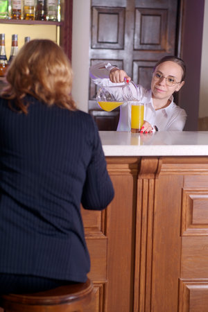 Young waitress in glasses pouring juice into glass from jug to unrecognizable woman, sitting at bar in restaurantの写真素材
