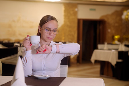 Young woman in glasses drinking coffee, tea, sitting in a restaurant, looking at watch on hand. Concept of time, busy, late, workload, employmentの写真素材