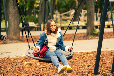 A beautiful and cheerful woman in warm clothes swings on a swing in the park. Girl swinging in the...