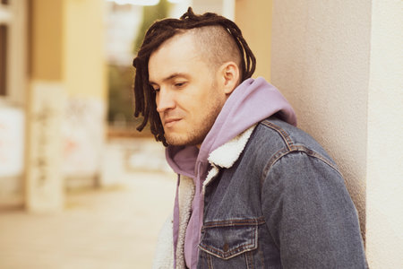 Portrait of young handsome man with dreadlocks posing, standing, leaning against wall on street