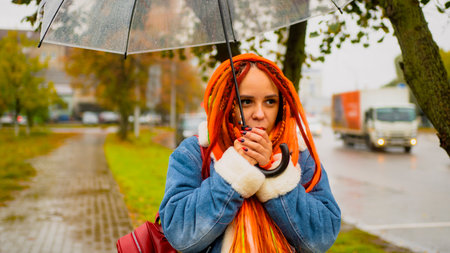 Young frozen woman with colorful dreadlocks, transparent umbrella standing in rain clenching hands...
