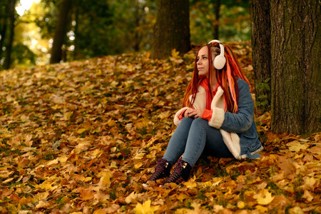 Young woman in headphones and listening to music sitting near tree in autumn forest.の写真素材
